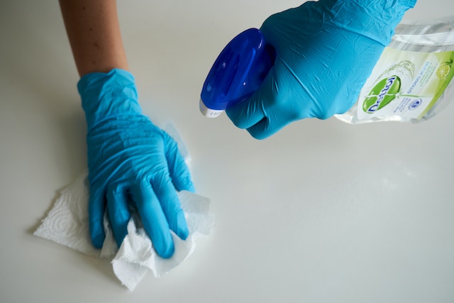 Blue-gloved hands spraying a bottle of cleaner and wiping a counter with a paper towel